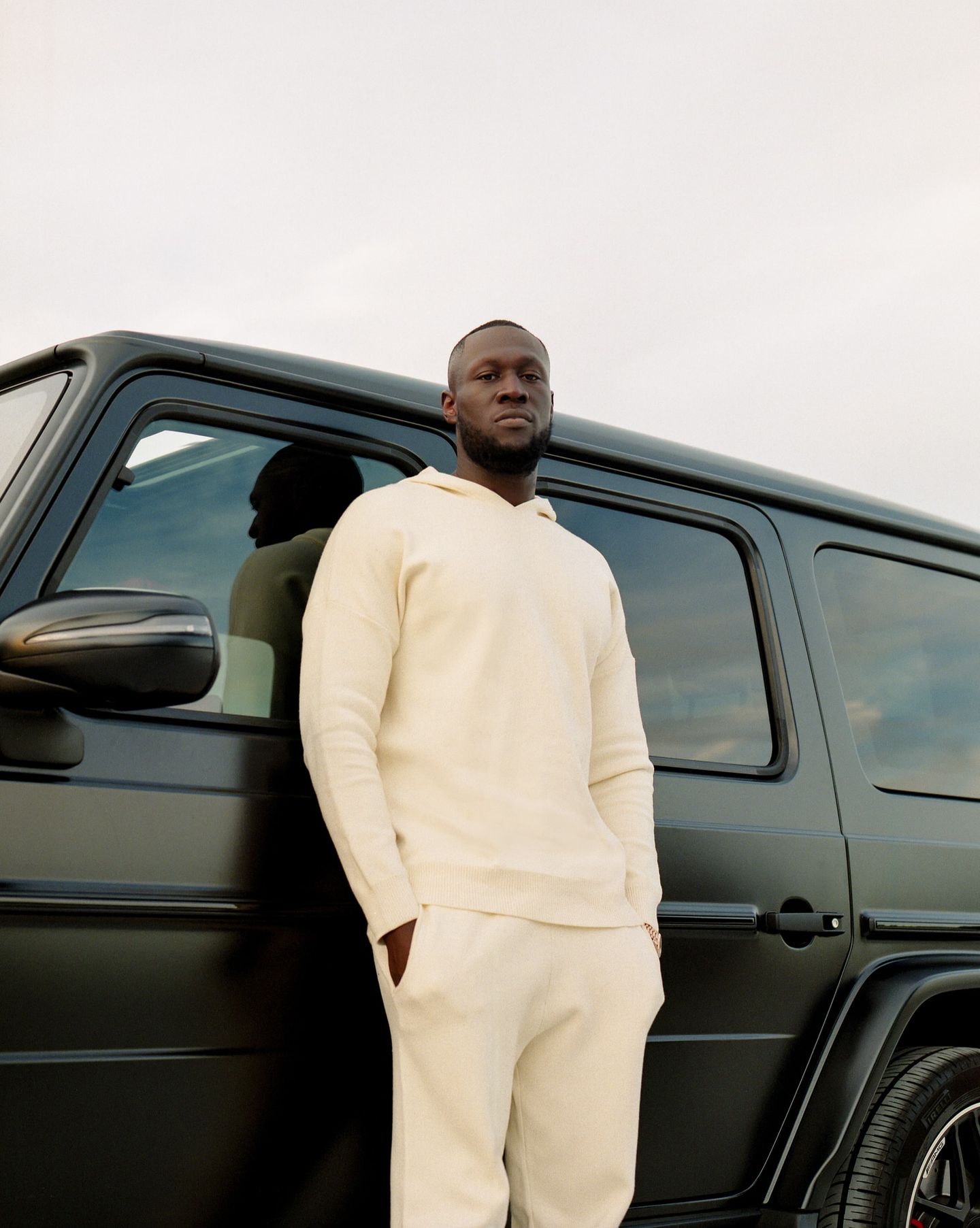 Stormzy in a cream outfit beside a dark vehicle